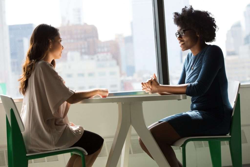 Two women discussing something while sitting at a modern table in a well-lit room in front of a window overlooking a large cityscape.
