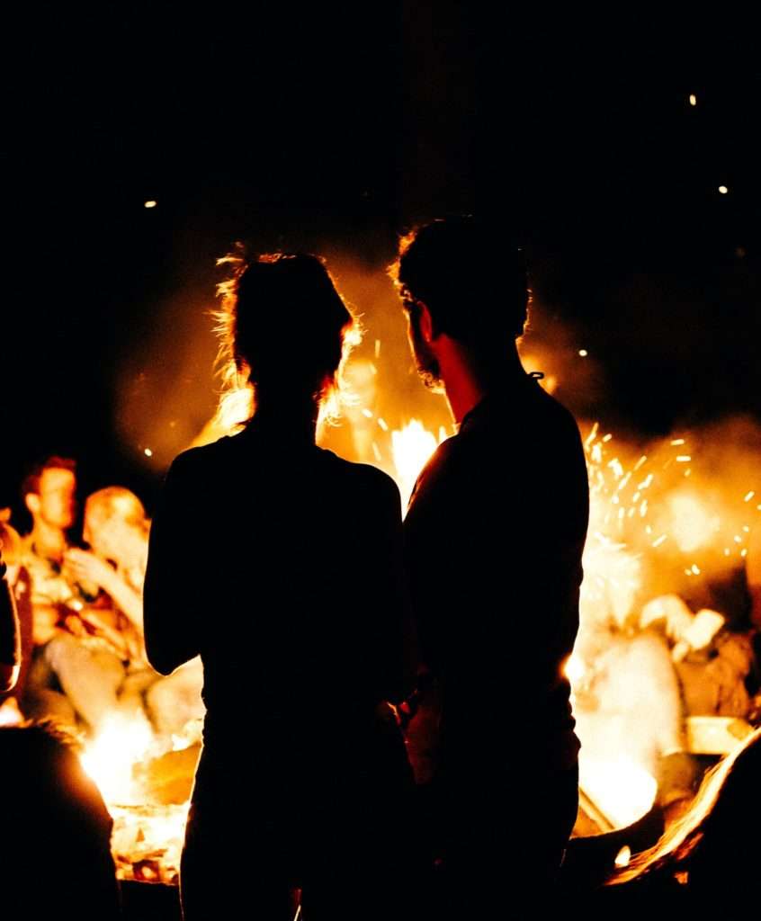 man and woman standing close to one another facing a fire at night in the dark.
